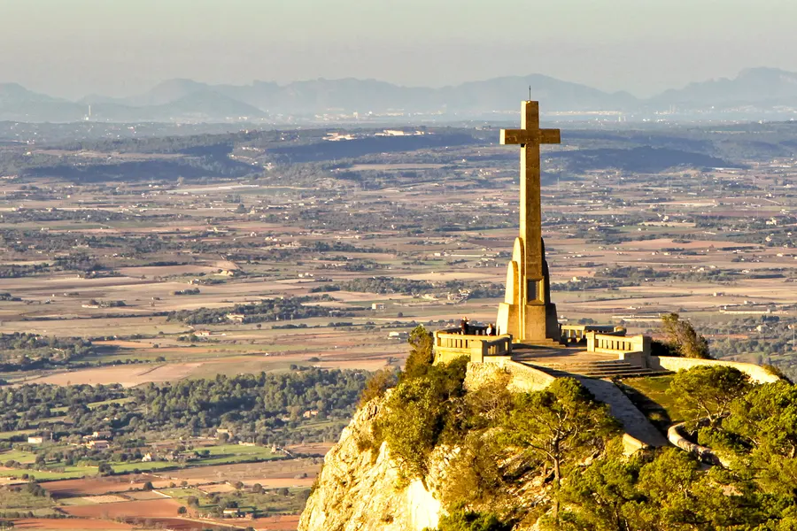 Großes steinernes Kreuz auf einer gemauerten Aussichtsplattform am Rand des Felssporns, dahinter die weite Ebene des Pla bis zu den Bergen der Tramuntana im Dunst.
