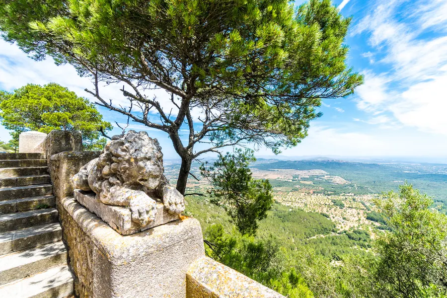 Steinerner Löwe auf einer Brüstung am Aufgang zum Kloster Sant Salvador, dahinter Kiefern und der Blick in die Ebene.
