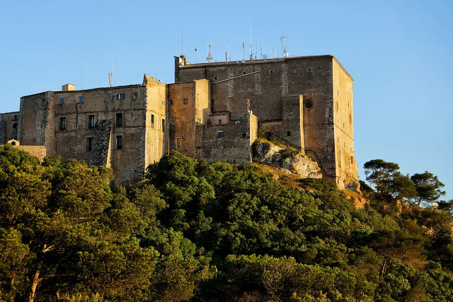 Der langgestreckte Klosterbau von Sant Salvador im Abendlicht auf dem Felsgrat, darunter dichter Steineichenwald.
