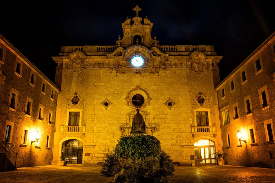 Die angestrahlte Fassade der Klosterkirche von Lluc bei Nacht, davor der leere Hof und die beleuchteten Arkaden.