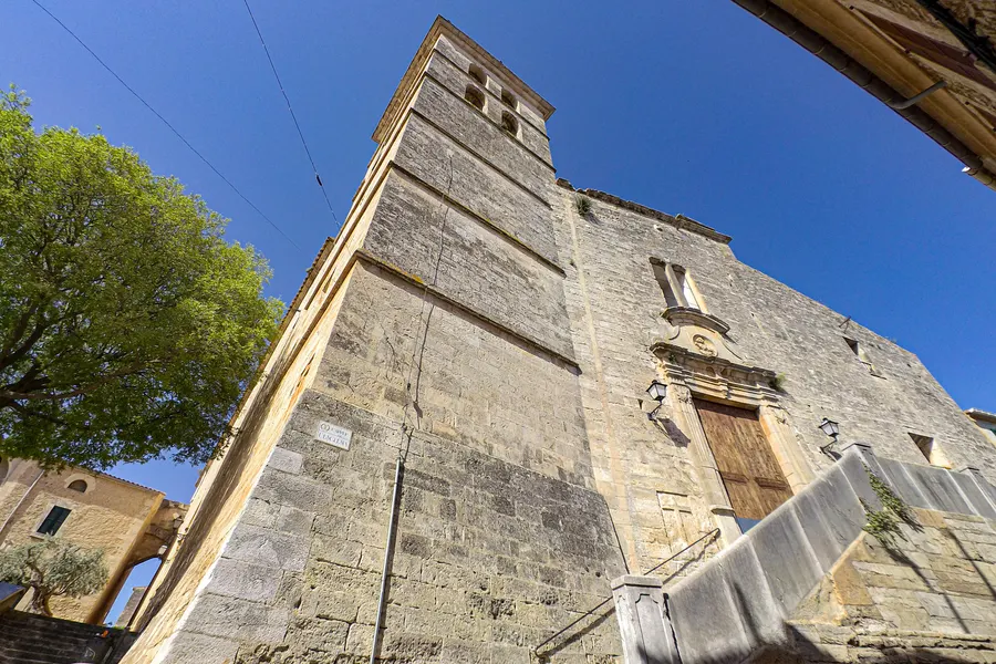 Die hohe schlichte Sandsteinfassade der Kirche von Sencelles von unten gesehen, mit schmalem Glockengiebel und Seitenportal.
