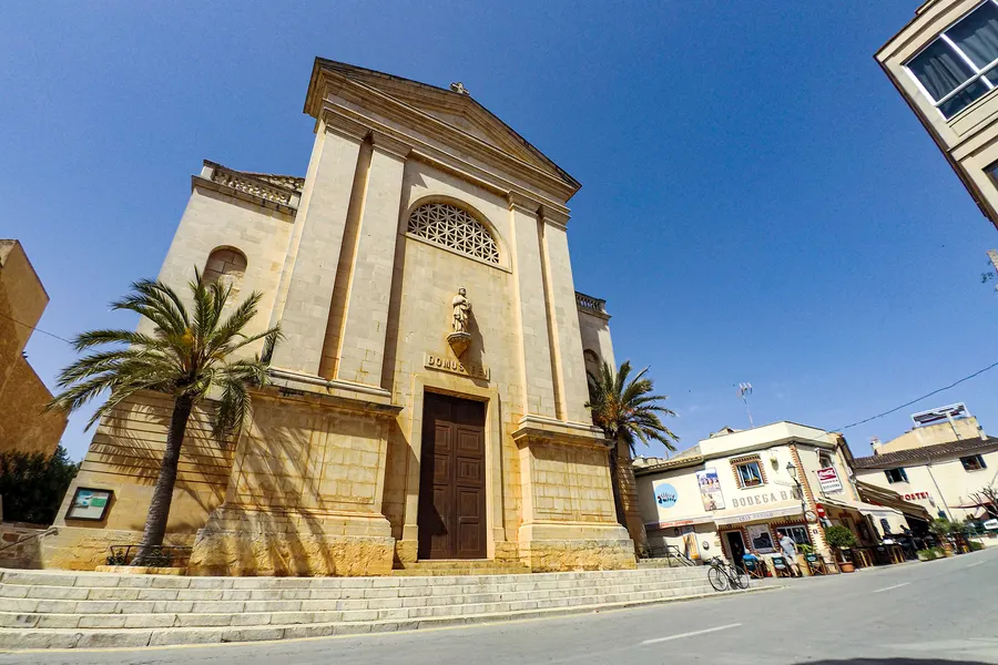 Die Kirche von Ses Salines mit heller Sandsteinfassade, Rosettenfenster und Freitreppe, davor eine hohe Palme und der Platz.
