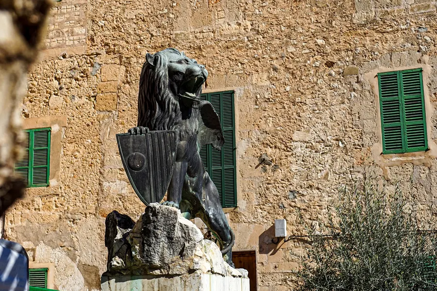 Der bronzene geflügelte Löwe von Sineu mit Wappenschild auf einem Steinsockel, dahinter eine Sandsteinfassade mit grünen Fensterläden und ein Olivenbaum.
