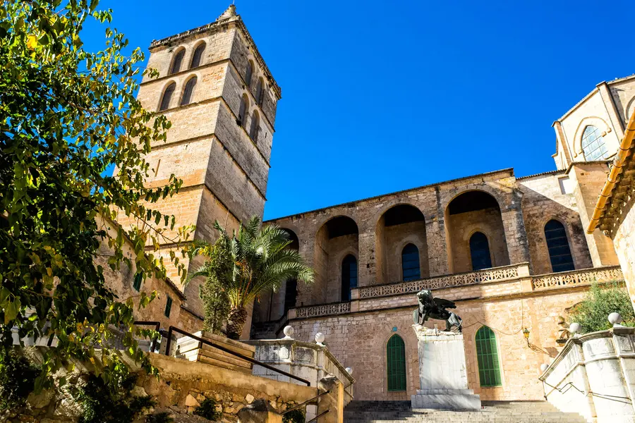 Der wuchtige Glockenturm der Kirche von Sineu und die Arkadengalerie an ihrer Seite, davor Palmen und eine Freitreppe.
