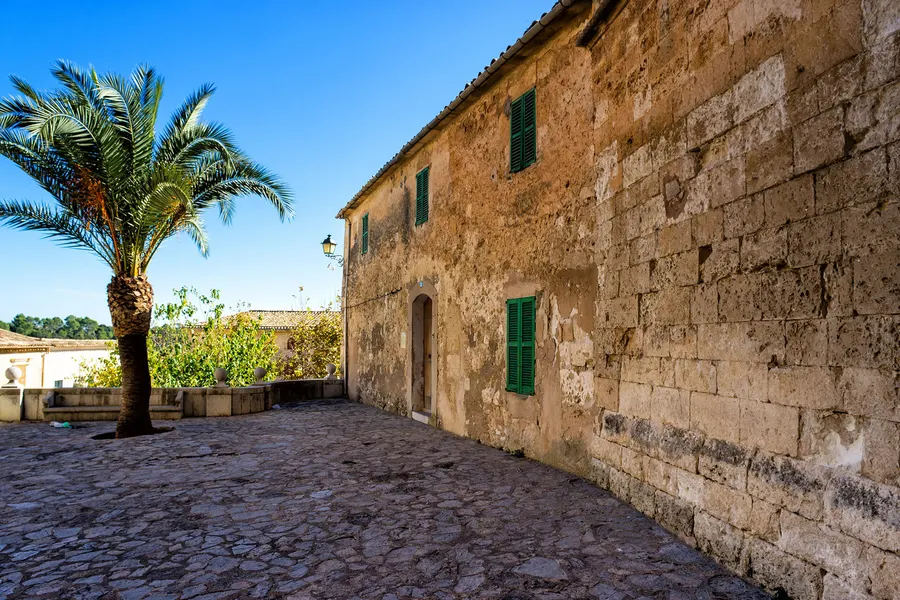 Lange fensterarme Sandsteinmauer entlang einer gepflasterten Gasse in Sineu, davor eine Palme unter blauem Himmel.
