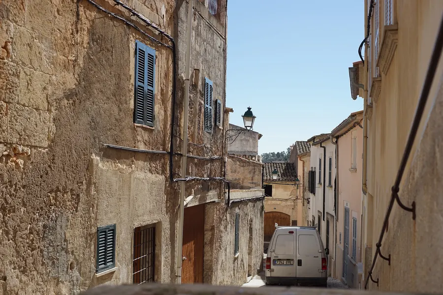 Enge Gasse zwischen hohen Sandsteinhäusern in Sineu mit blauen Fensterläden und einem geparkten Lieferwagen.
