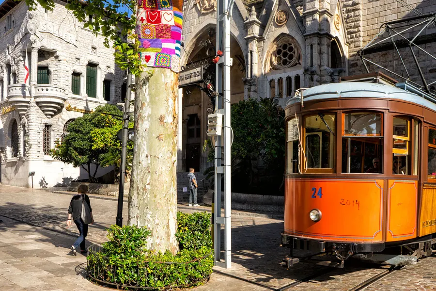 Die orangefarbene Straßenbahn von Sóller auf ihren Gleisen am Hauptplatz, dahinter die Kirche und ein bemalter Baumstamm.