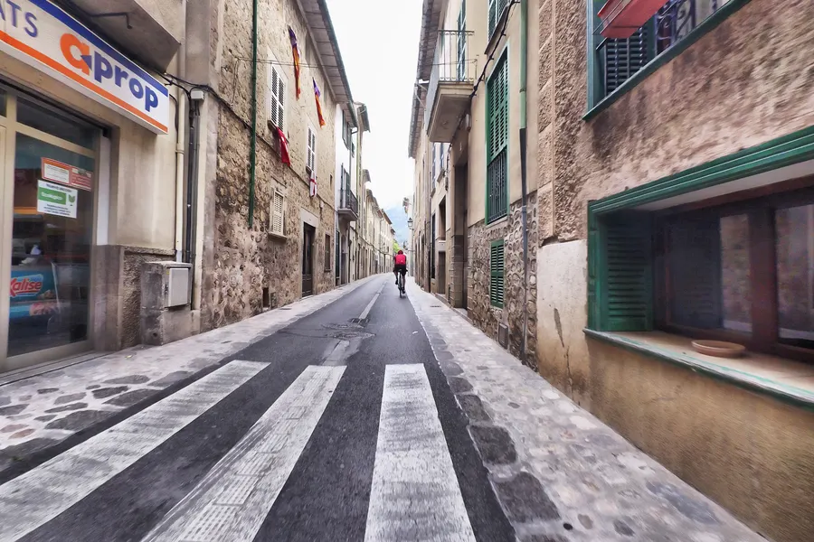 Straße in Sóller mit Zebrastreifen und einem Radfahrer in der Ferne, links ein Immobilienbüro, rechts Natursteinfassaden.