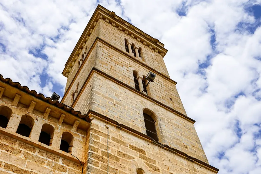 Der eckige Glockenturm der Kirche Sant Miquel von unten fotografiert, mit Schallöffnungen und Zinnenkranz vor Wolkenhimmel.
