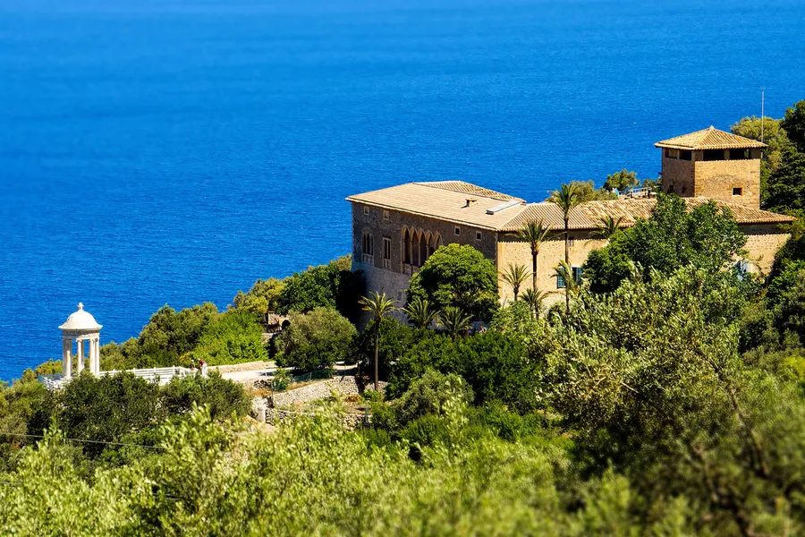 Das Herrenhaus Son Marroig aus hellem Naturstein mit Turm auf einer bewaldeten Terrasse hoch über dem Meer, davor der weiße Rundtempel.