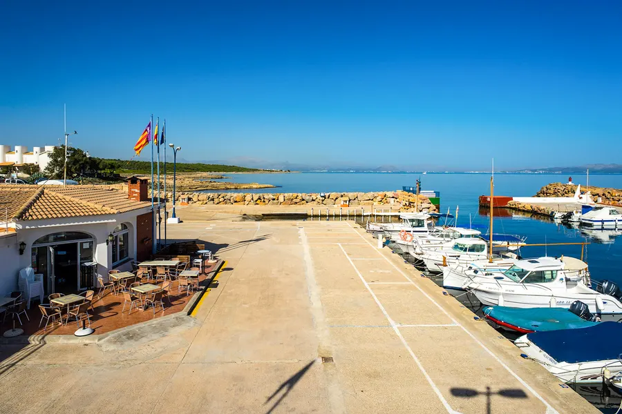 Weite betonierte Hafenfläche mit einem flachen Lokal mit Terrasse und Fahnen, rechts liegende Boote, dahinter das offene Meer.