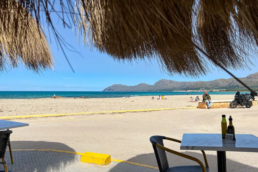Blick aus einer Strandbar unter einem Schilfdach hinaus auf den Strand von Son Serra de Marina, im Hintergrund die Berge des Massís d'Artà.