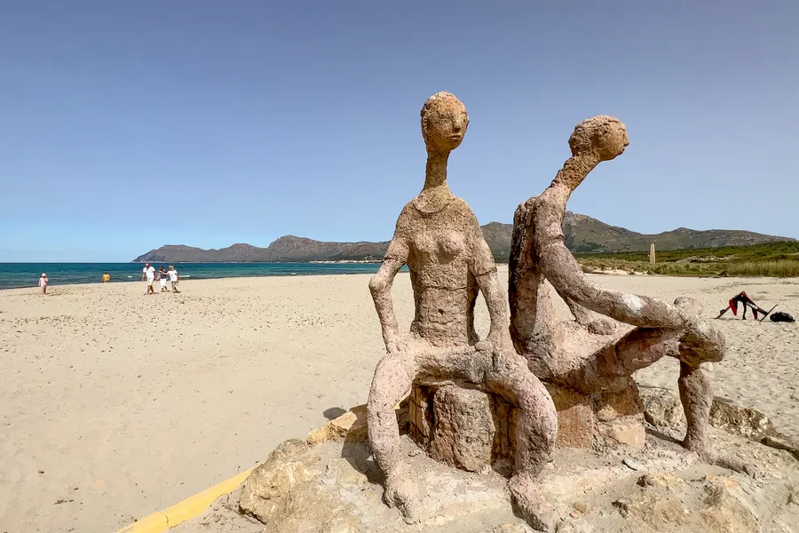 Steinerne Figurengruppe aus zwei sitzenden Gestalten auf einem Sockel am Strandrand von Son Serra de Marina, dahinter Sand, Wasser und Berge.
