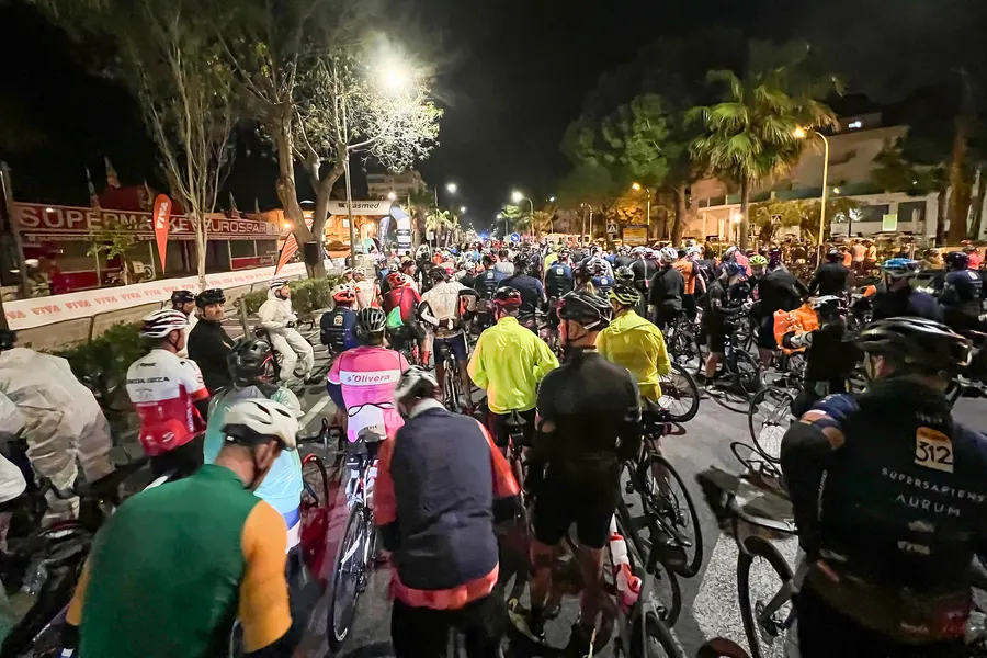Rennradfahrer im Dunkeln am Start der Mallorca 312 in Platja de Muro, angestrahlt von Scheinwerfern und Startbogen.