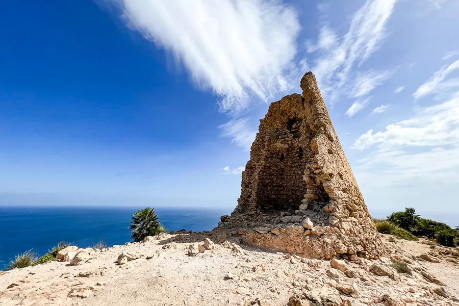 Abgebrochener Mauerrest des Turms als spitze Silhouette gegen einen blauen Himmel mit Federwolken, unten das Meer.