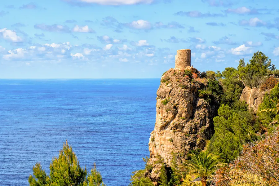Der Torre des Verger auf seinem Felsen über dem Meer, im Vordergrund Kiefern und niedriger Bewuchs unter Schleierwolken.