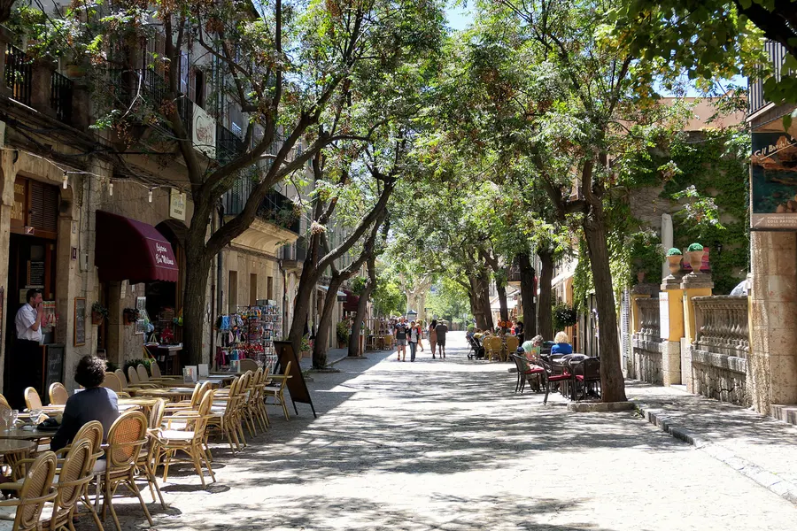 Baumbestandene Straße in Valldemossa mit Caféterrassen, Souvenirständen und Passanten, dahinter Häuser und Grün.