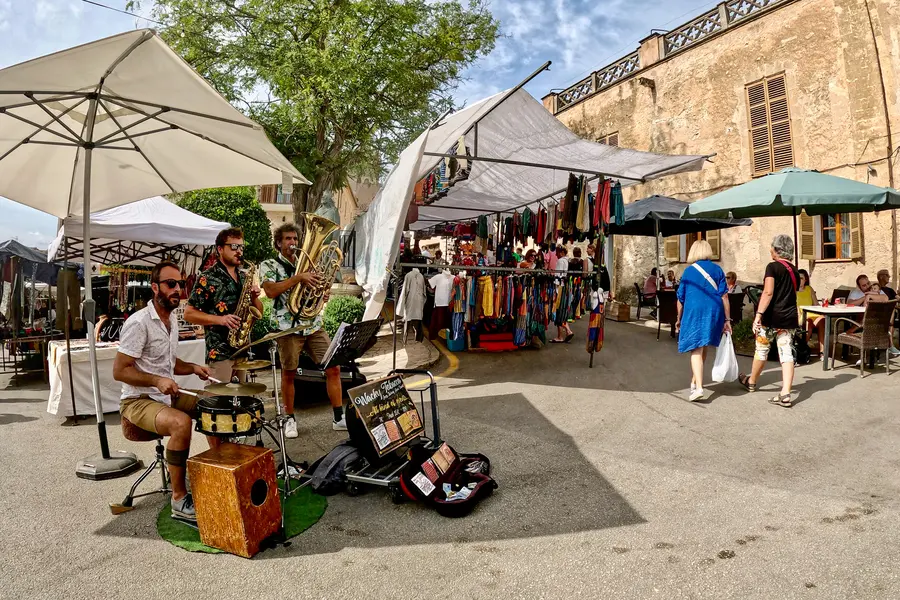 Marktstände mit weißen Sonnenschirmen auf einem gepflasterten Platz in Sineu, im Schatten alter Bäume, davor ein Straßenmusiker mit Instrumenten.