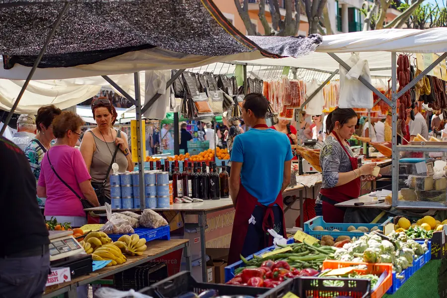 Gemüsestand auf dem Markt von Alcúdia mit Kisten voller Obst und Gemüse, davor mehrere Kundinnen und eine Verkäuferin.